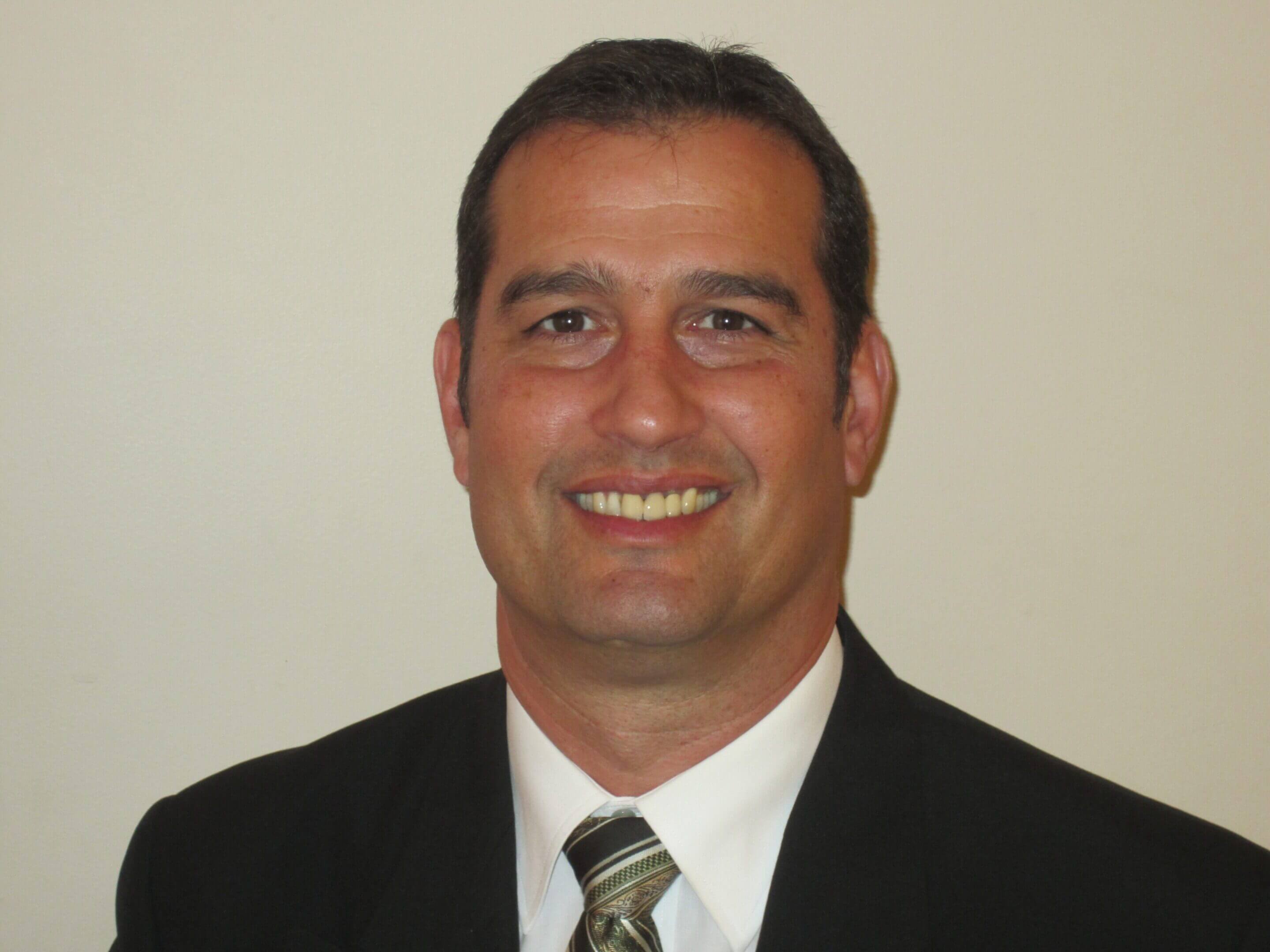 Smiling man in a suit with a striped tie against a plain background.