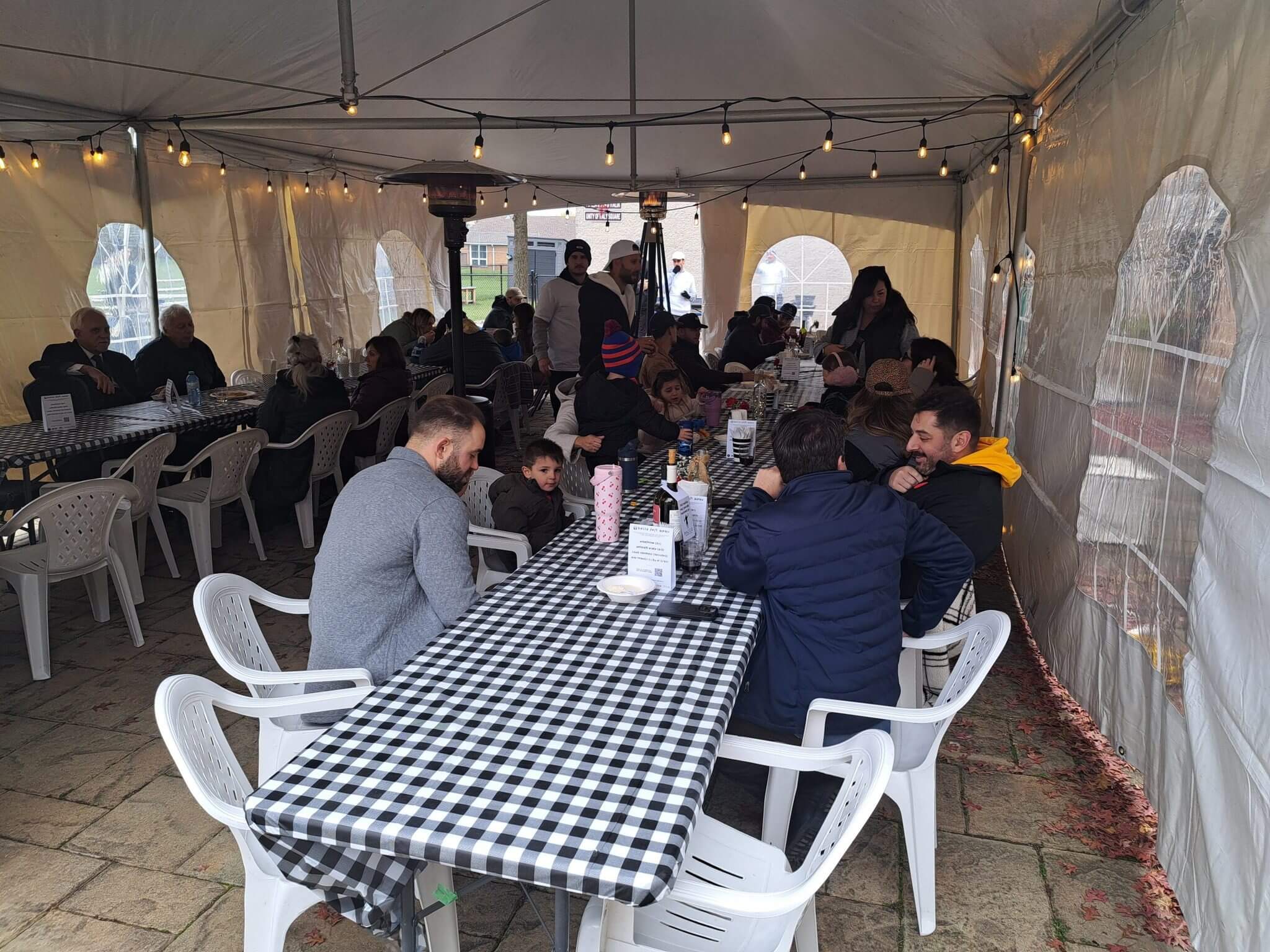 People gathered under a tent, sitting at tables with checkered tablecloths.
