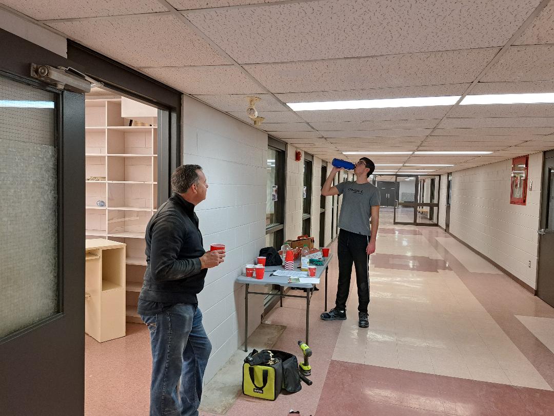 Two men in a hallway with a table of items between them.