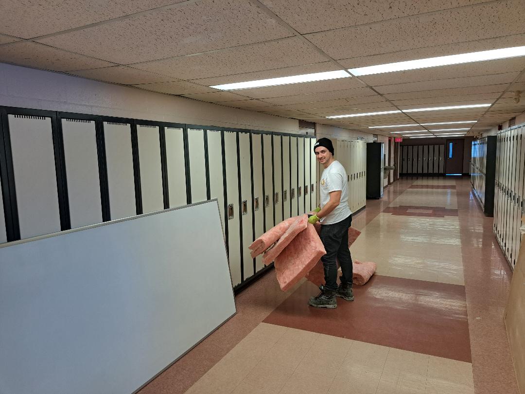 Person moving large boards and a rolled-up carpet in a hallway.
