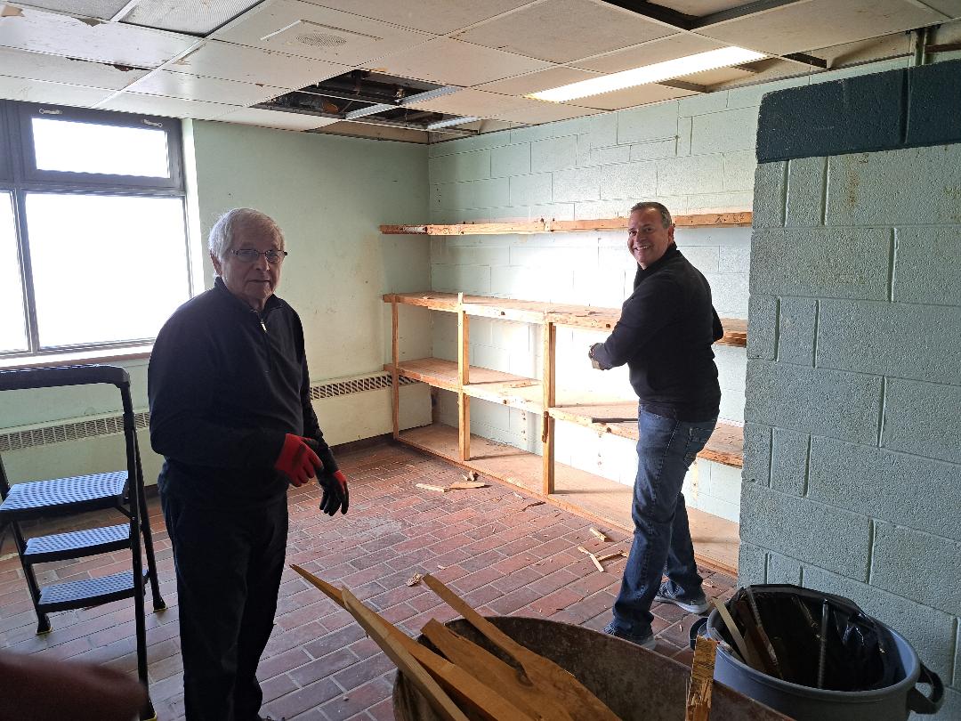 Two men working in a room with wall damage and empty shelves.