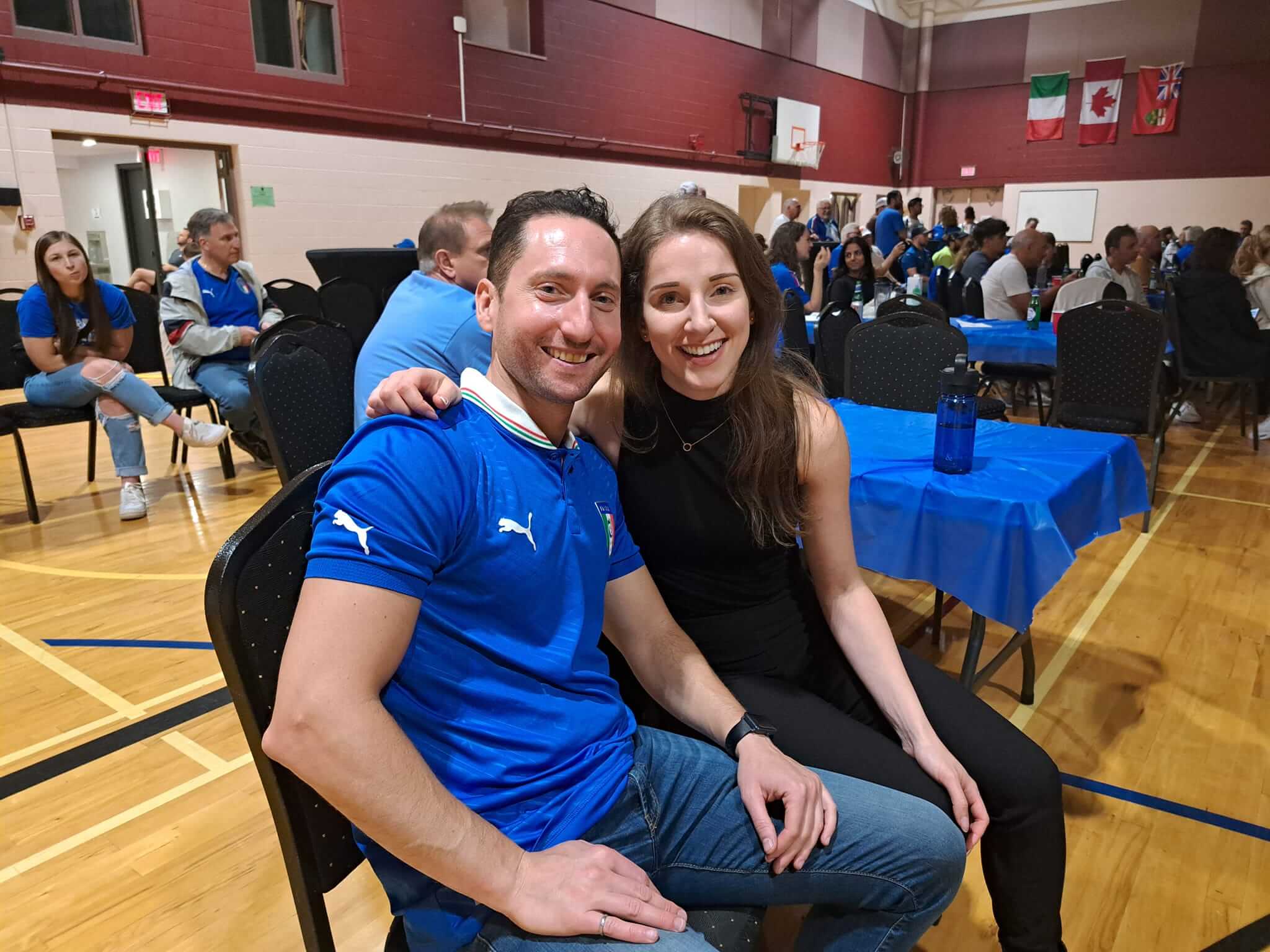 A smiling couple seated closely at a social event in a gymnasium.