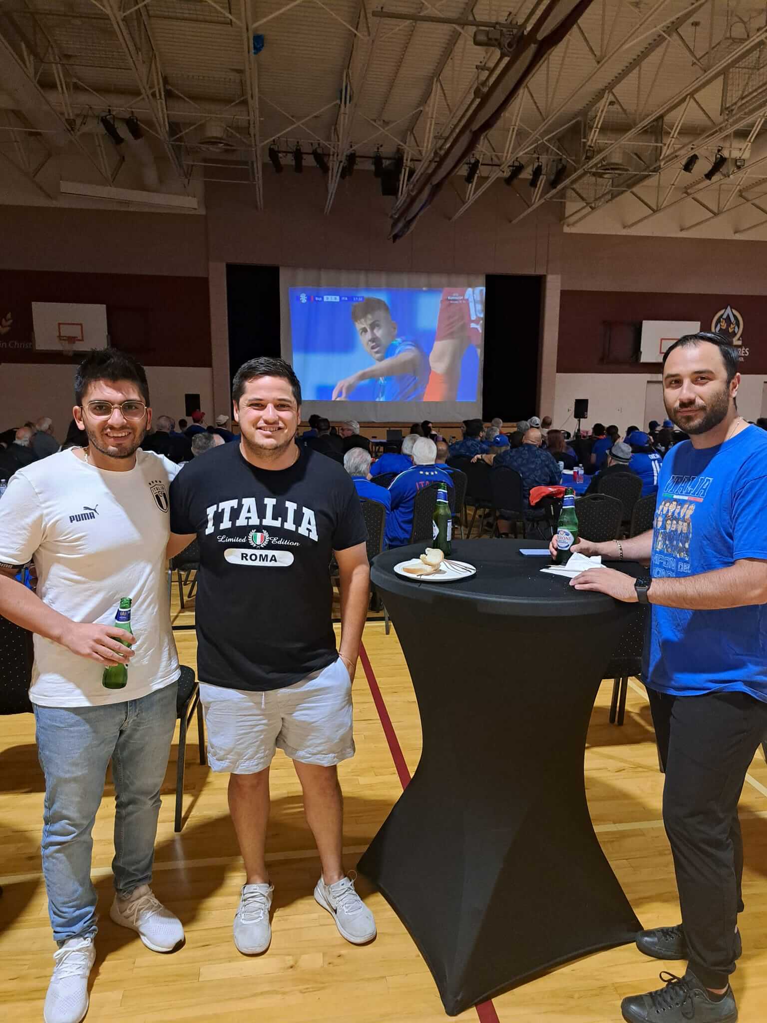 Three men posing casually around a cocktail table with a sports game on a screen behind.