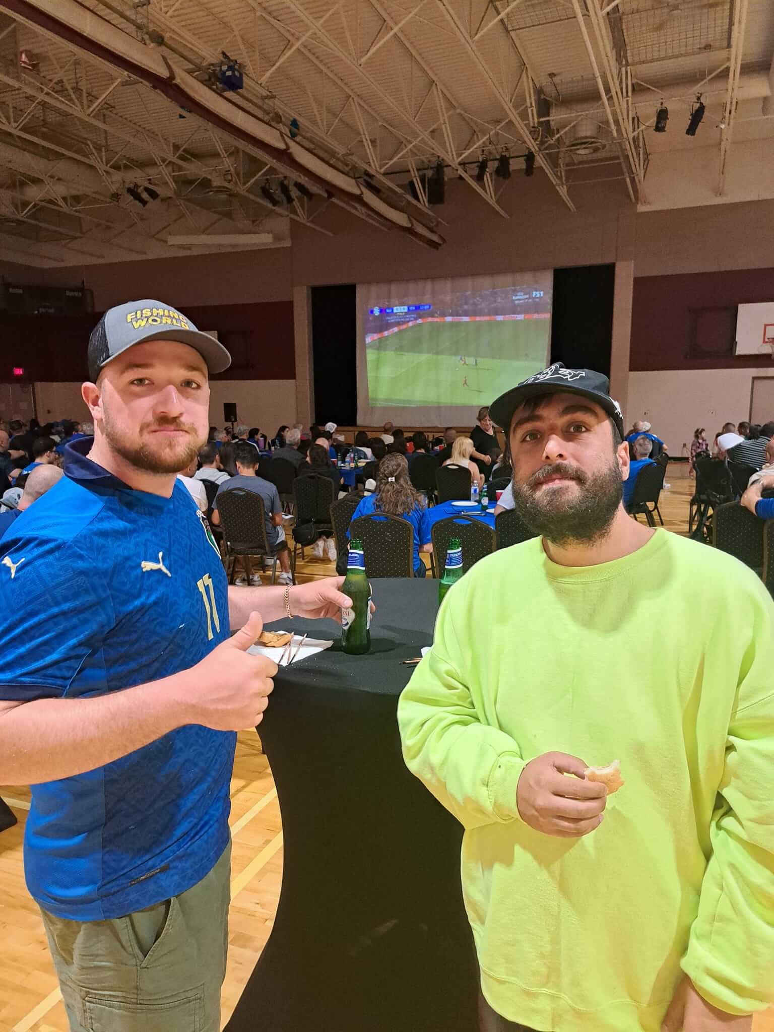 Two men posing together inside a crowded indoor venue with a large screen.