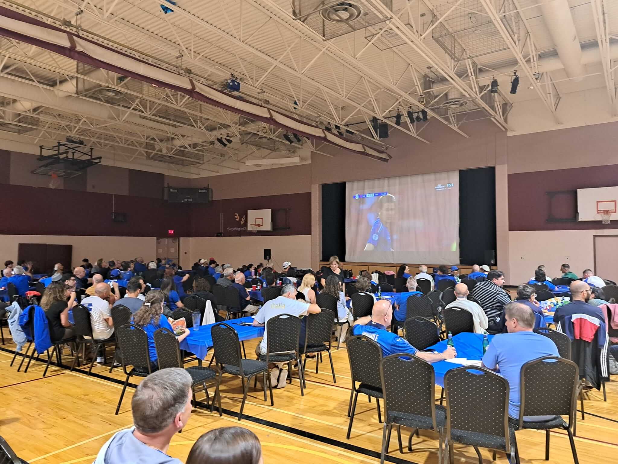 People attending a presentation in a gymnasium with a large screen.