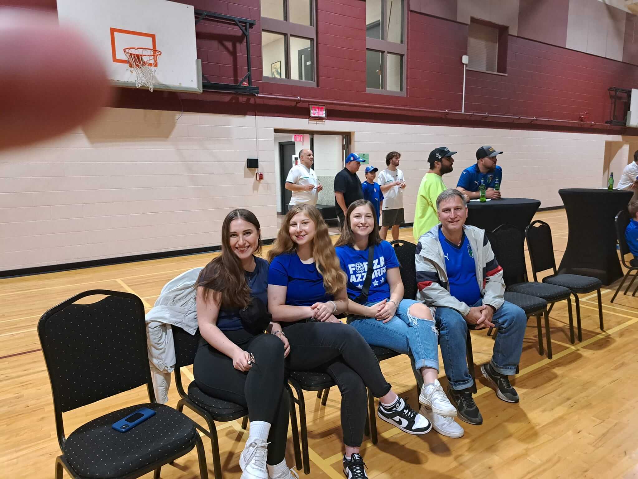 Group of smiling people seated in a gymnasium with basketball hoops in the background.