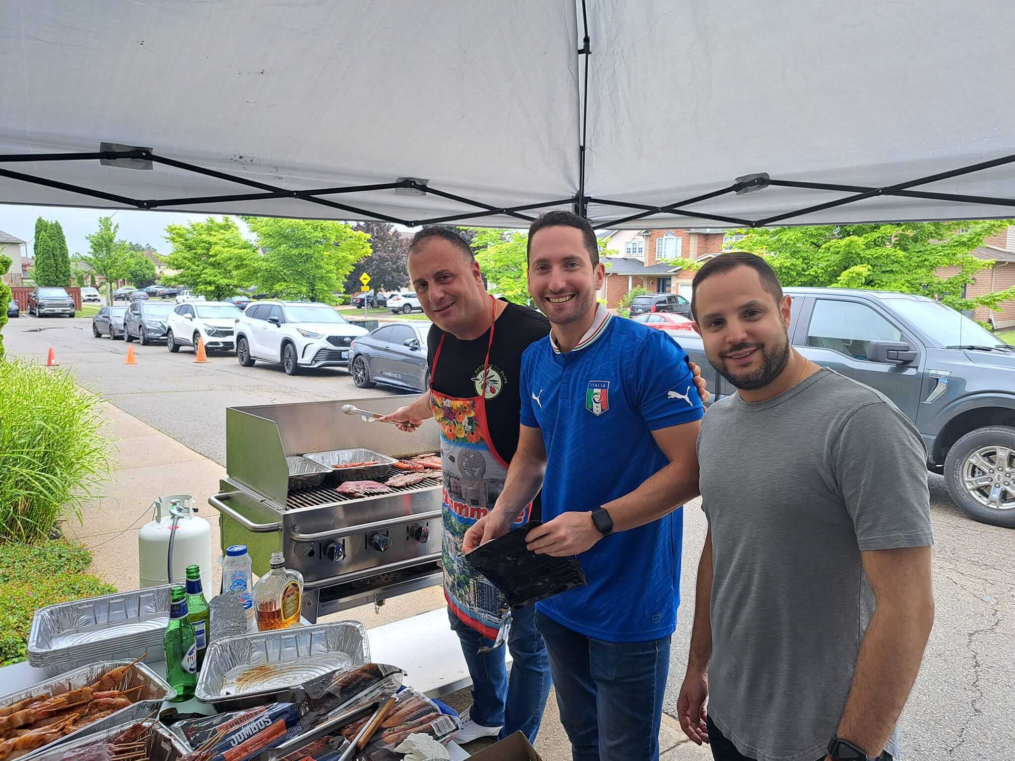 Three men smiling and cooking outdoors under a tent.