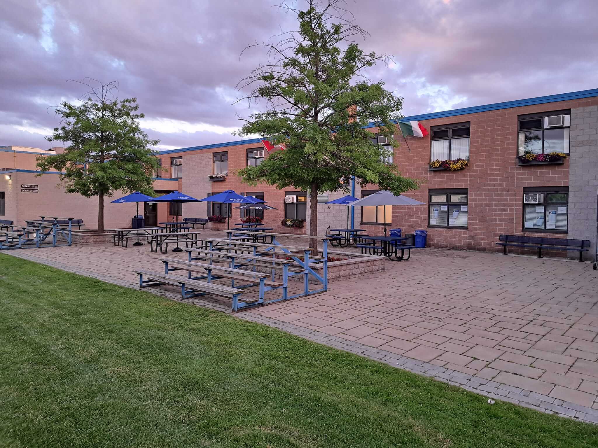 Outdoor seating area with blue umbrellas and picnic tables beside a grassy lawn.
