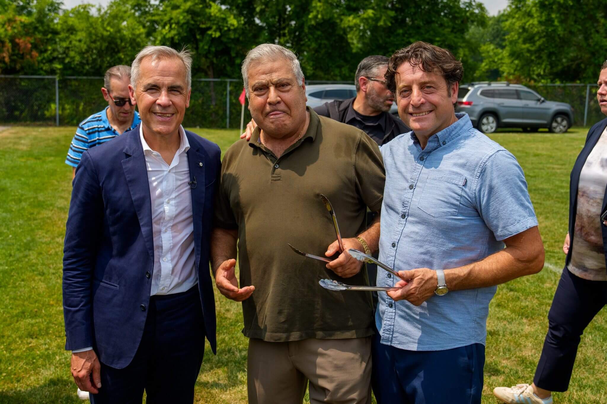 Three men smiling outdoors, holding glasses with greenery in the background.