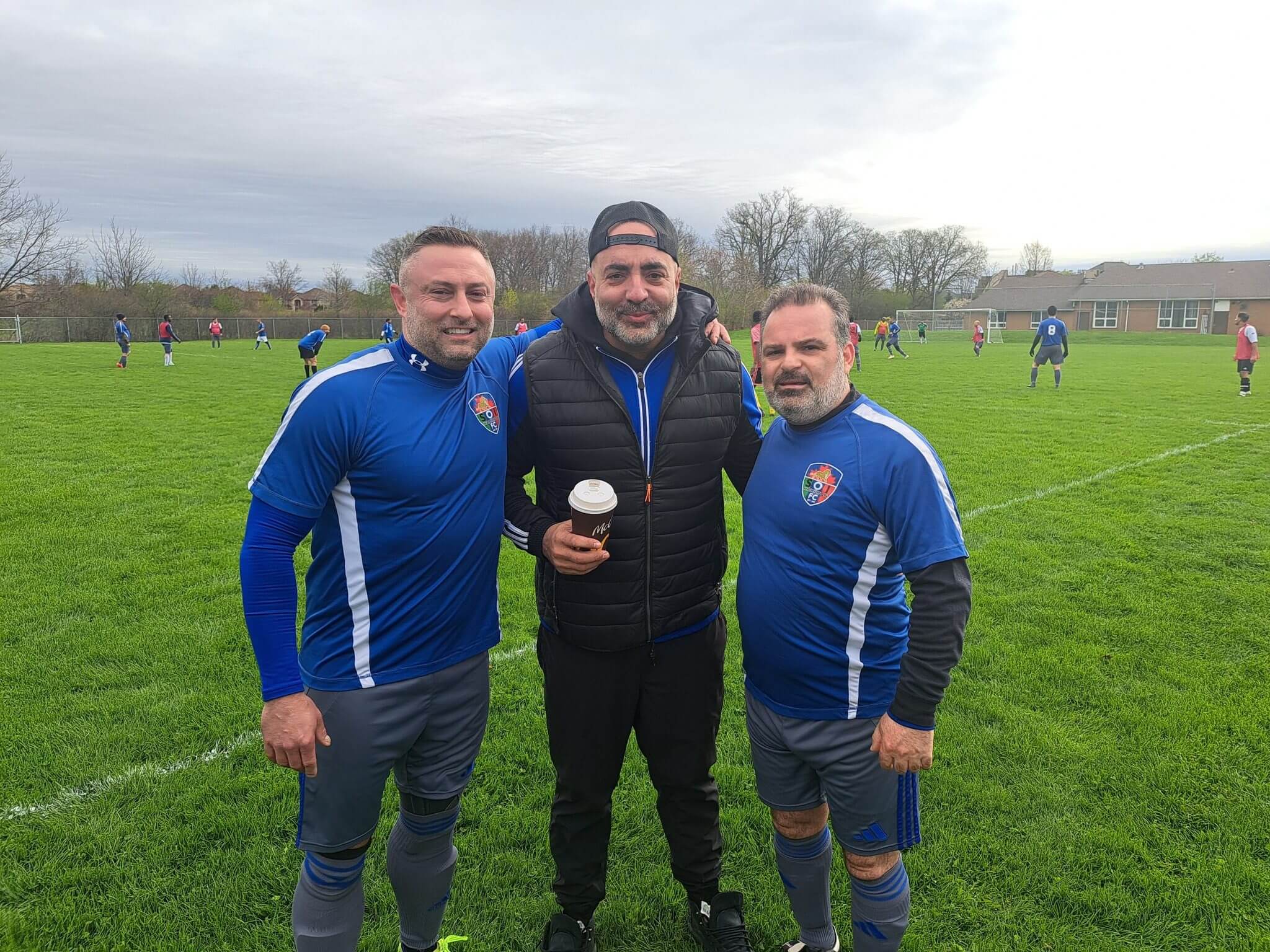 Three men posing on a grassy sports field, two in matching blue athletic gear.