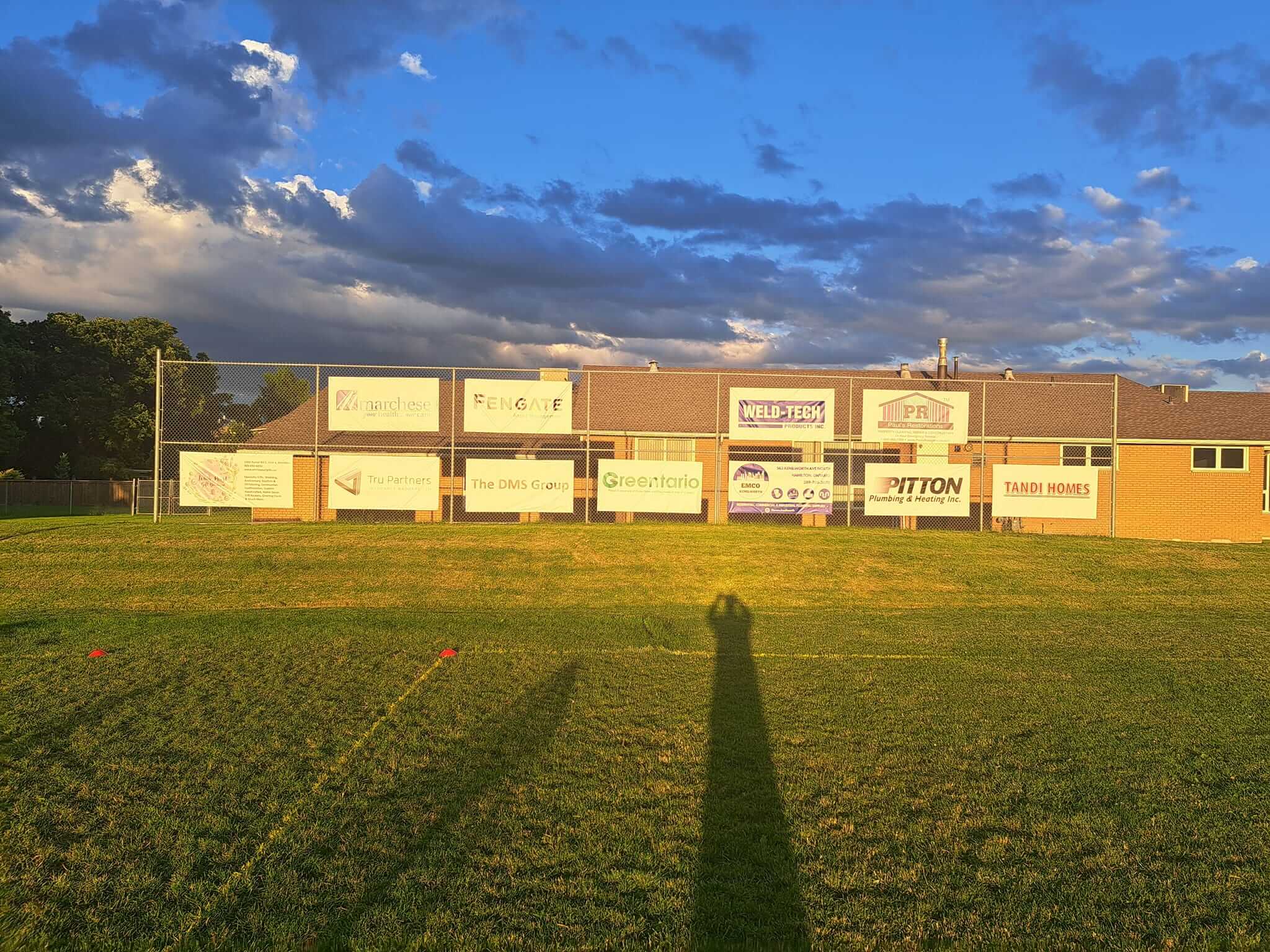 Sunset shadows stretch across a green field with a brick wall displaying various advertisements.