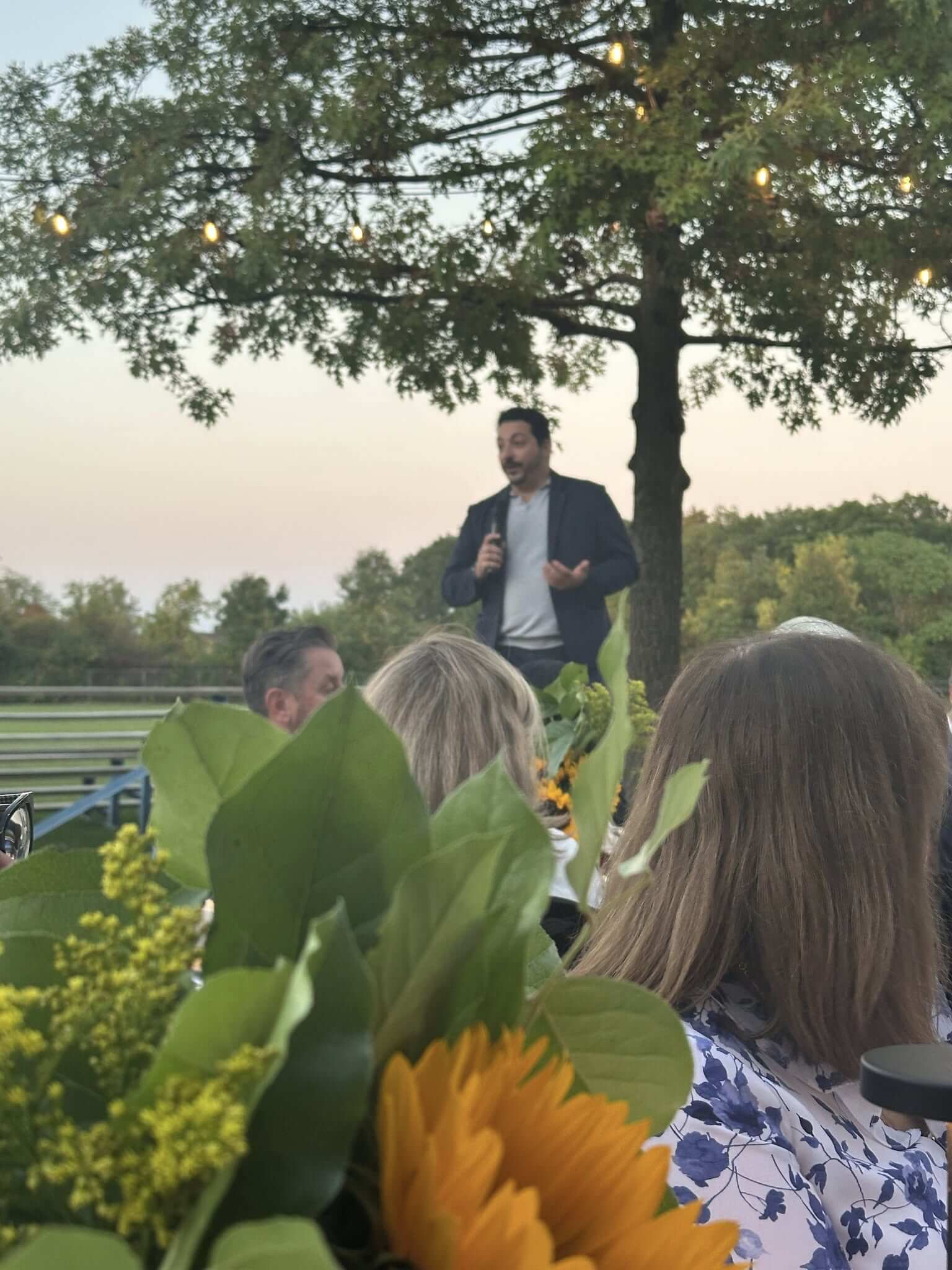 Man speaking to a crowd outdoors near a large tree at sunset.
