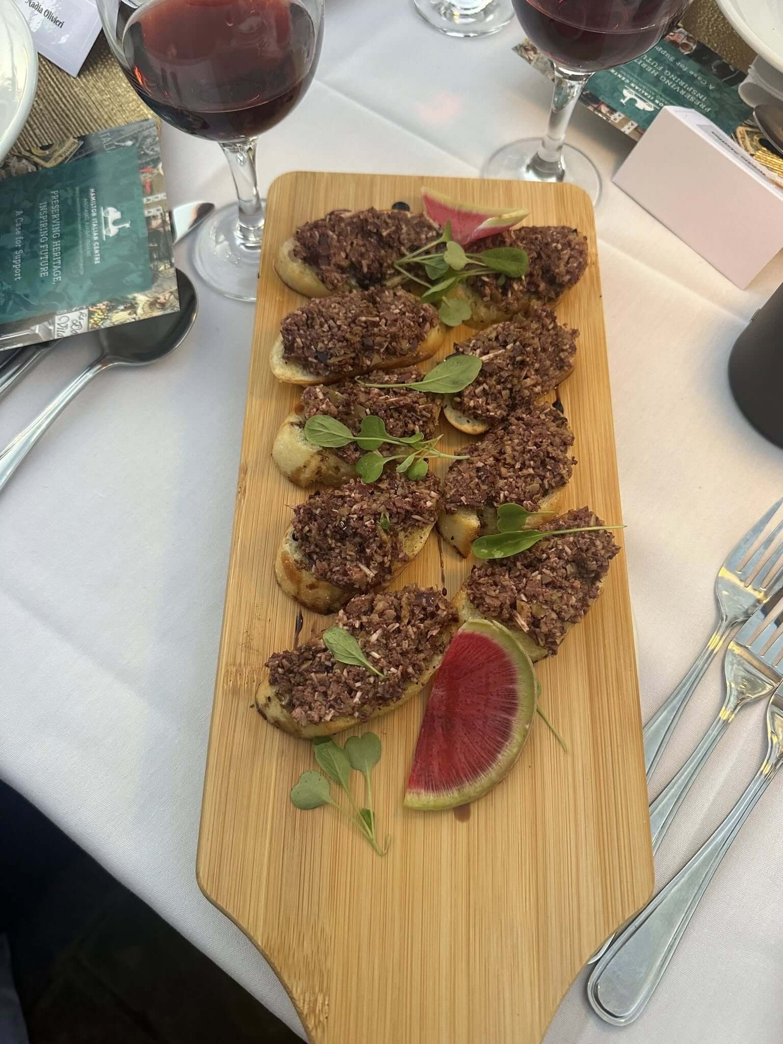 A wooden board with six servings of minced meat on flatbread garnished with herbs and vegetables.