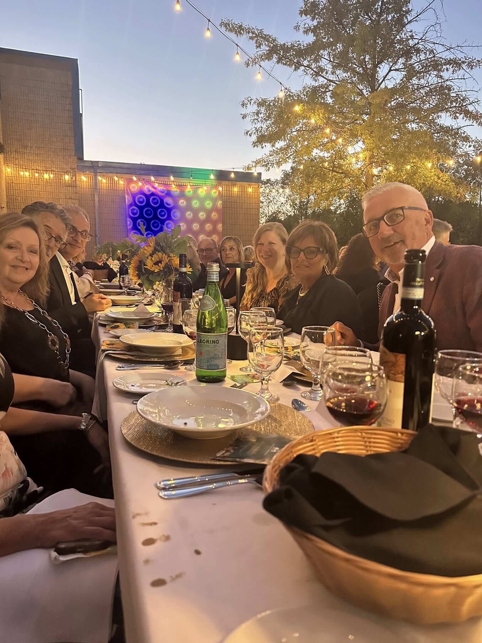 Friends enjoying a festive outdoor dinner with wine and string lights.