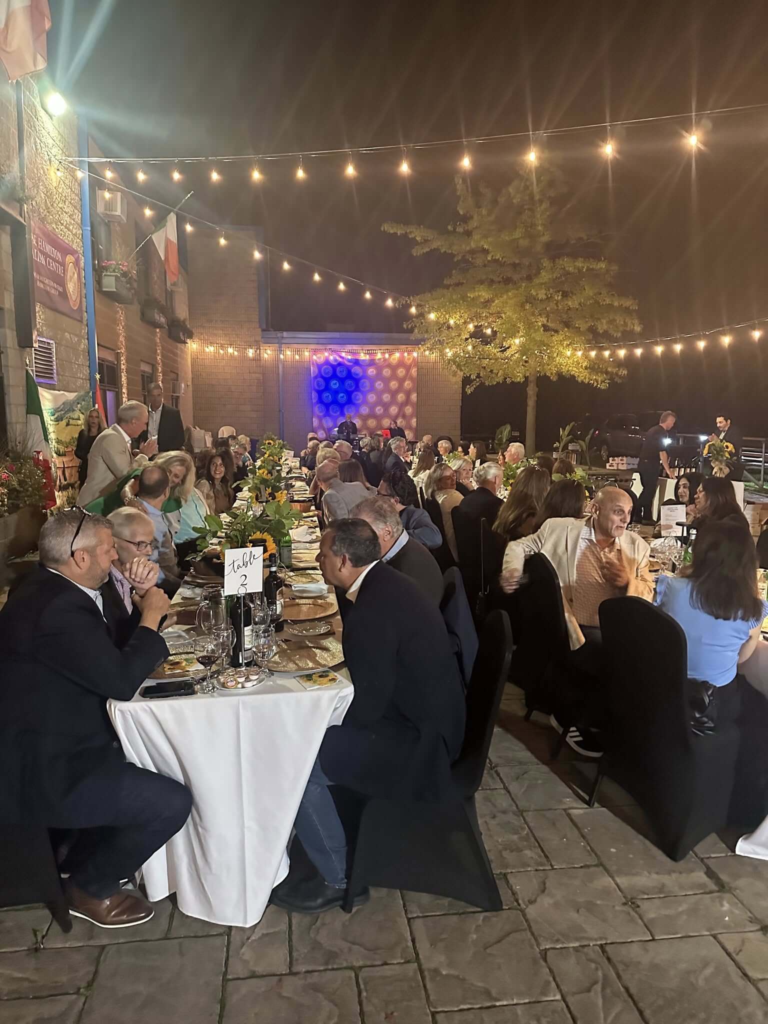 Elegant outdoor evening event with guests seated at round tables under string lights.