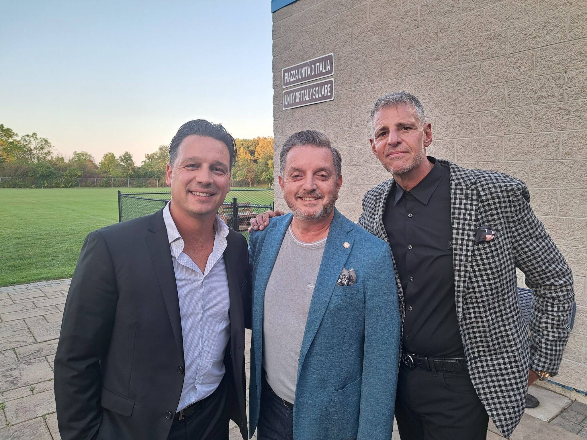 Three men posing together outdoors near a building at sunset.