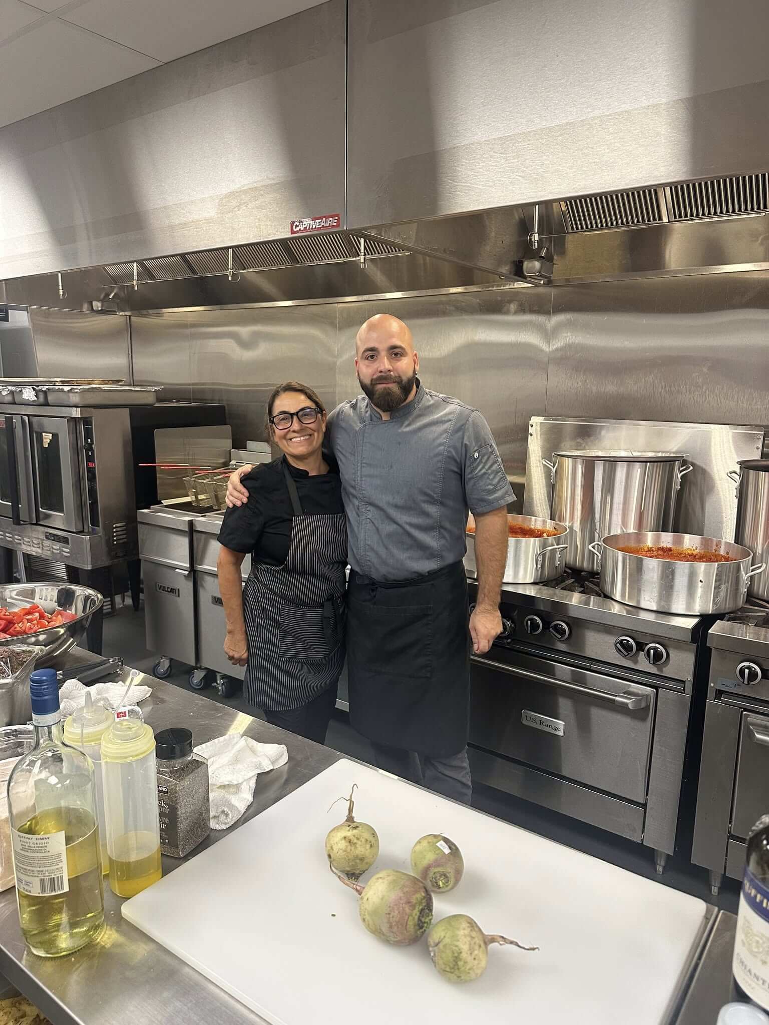 Two chefs posing in a professional kitchen with stainless steel appliances.