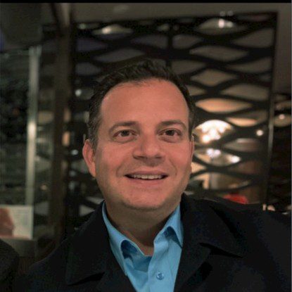 Smiling man in a blue shirt and dark jacket indoors.
