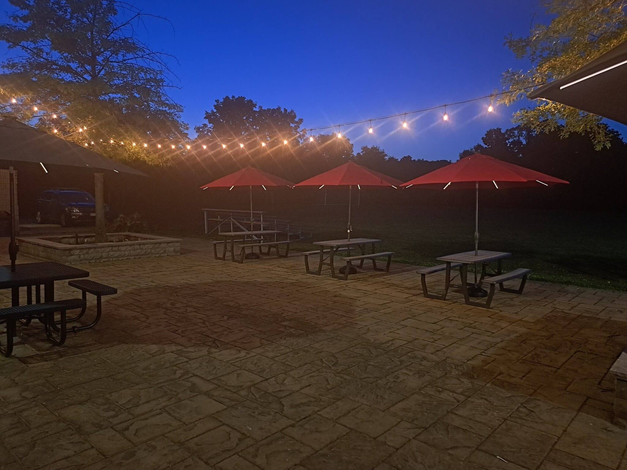 Outdoor seating area with red umbrellas and string lights at dusk.