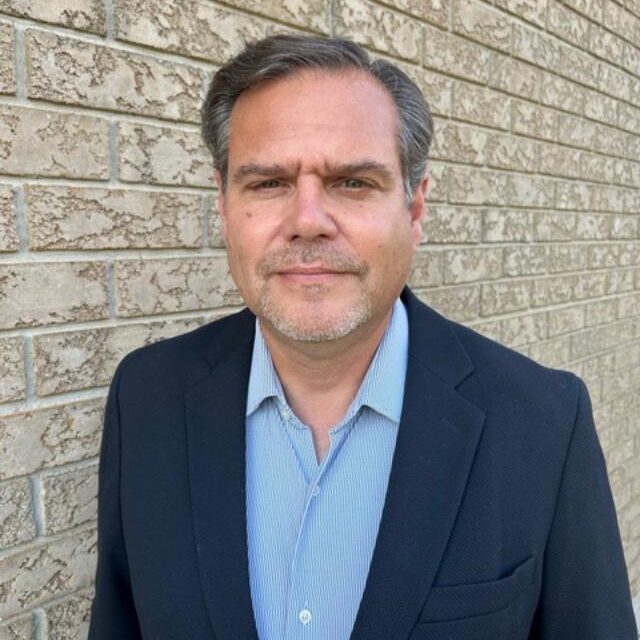 Man in a dark suit and blue shirt standing against a brick wall.
